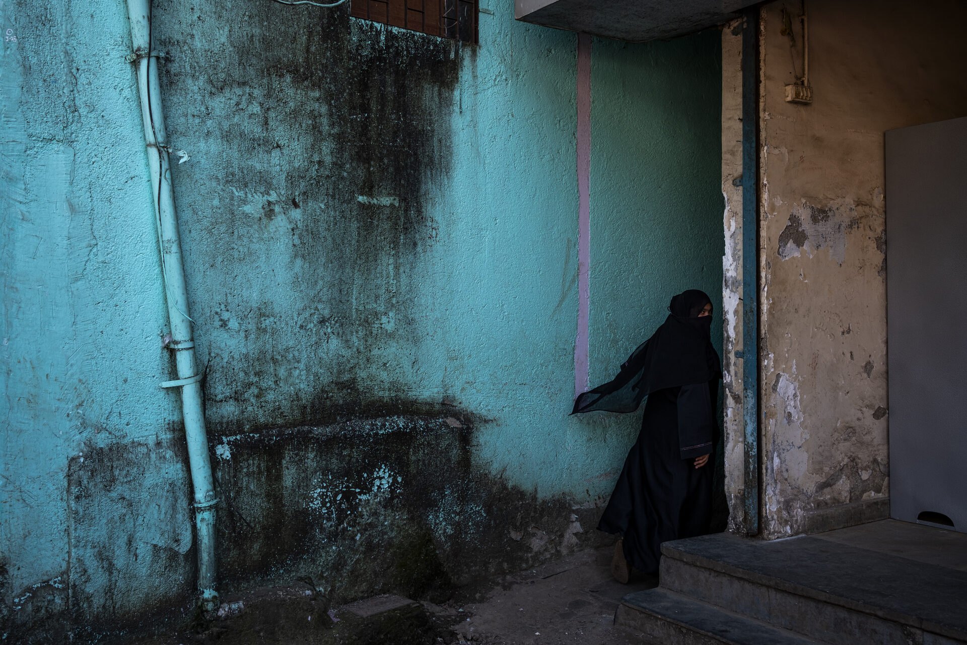 Mulher em niqab preto a passar por parede turquesa num bairro de Mumbai, Índia