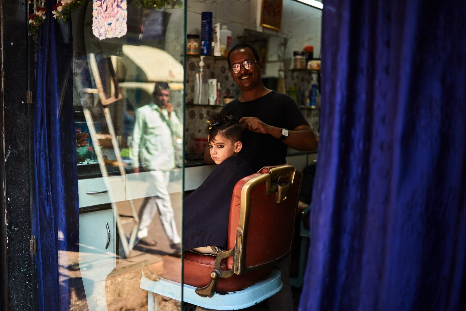 Menina a ser penteada num barbeiro de rua em Mumbai, vista por uma janela com cortinas azuis